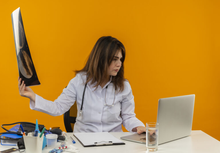 thinking middle aged female doctor wearing medical robe with stethoscope sitting at desk work on laptop with medical tools holding x ray and looking at laptop on isolated orange background with copy space
